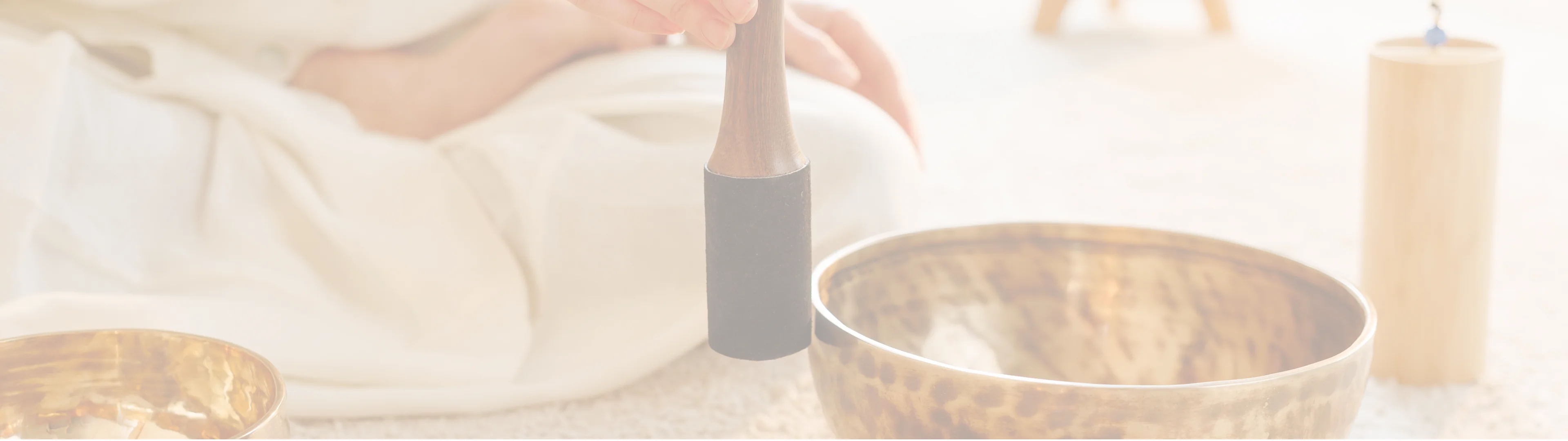 A softly lit scene showing a person wrapped in white fabric, holding a singing bowl set with a metallic bowl, a dark cylindrical object, and a candle.
