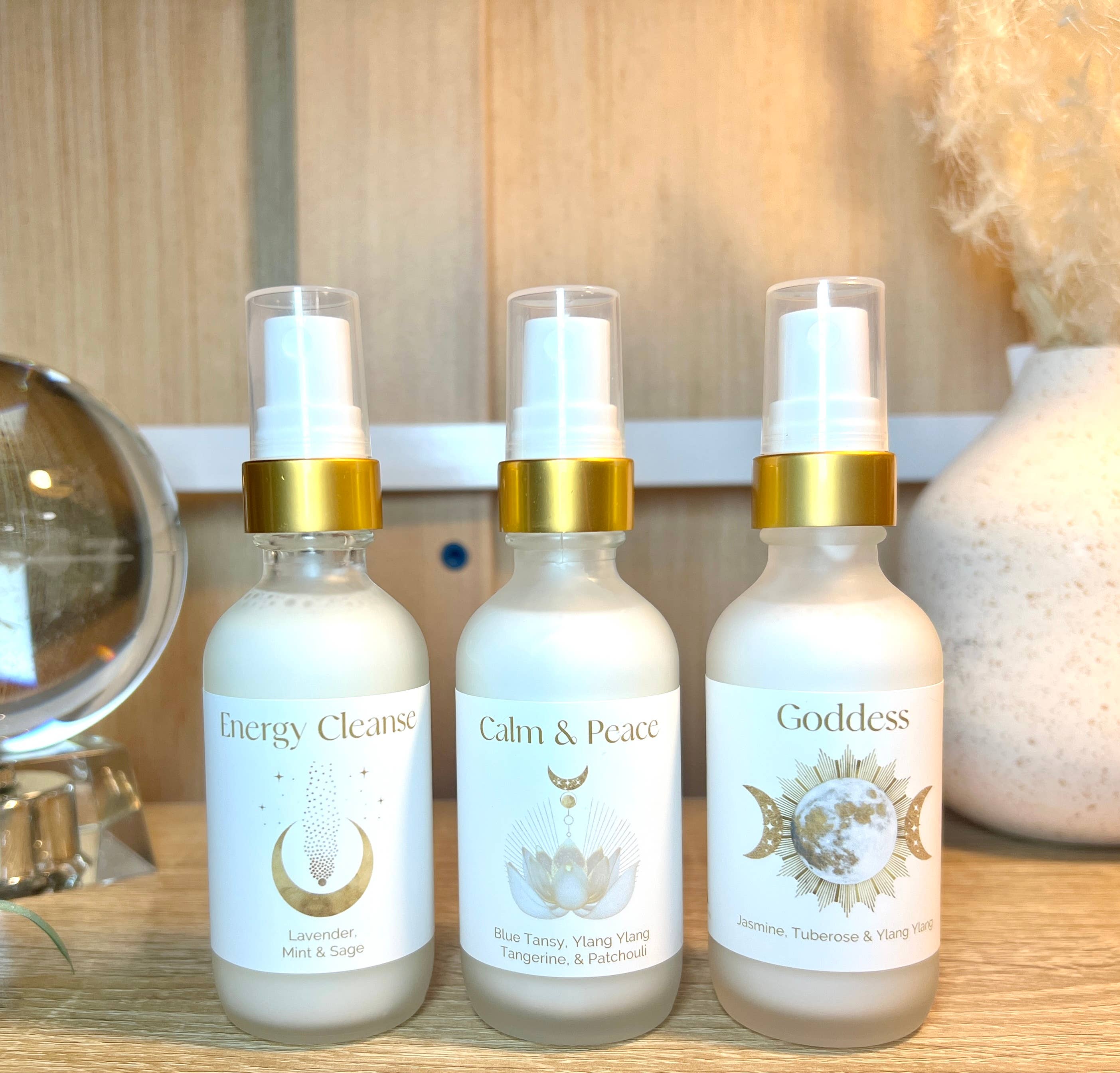 Three bottles labeled 'Energy Cleanse', 'Calm & Peace', and 'Goddess' on a wooden surface.