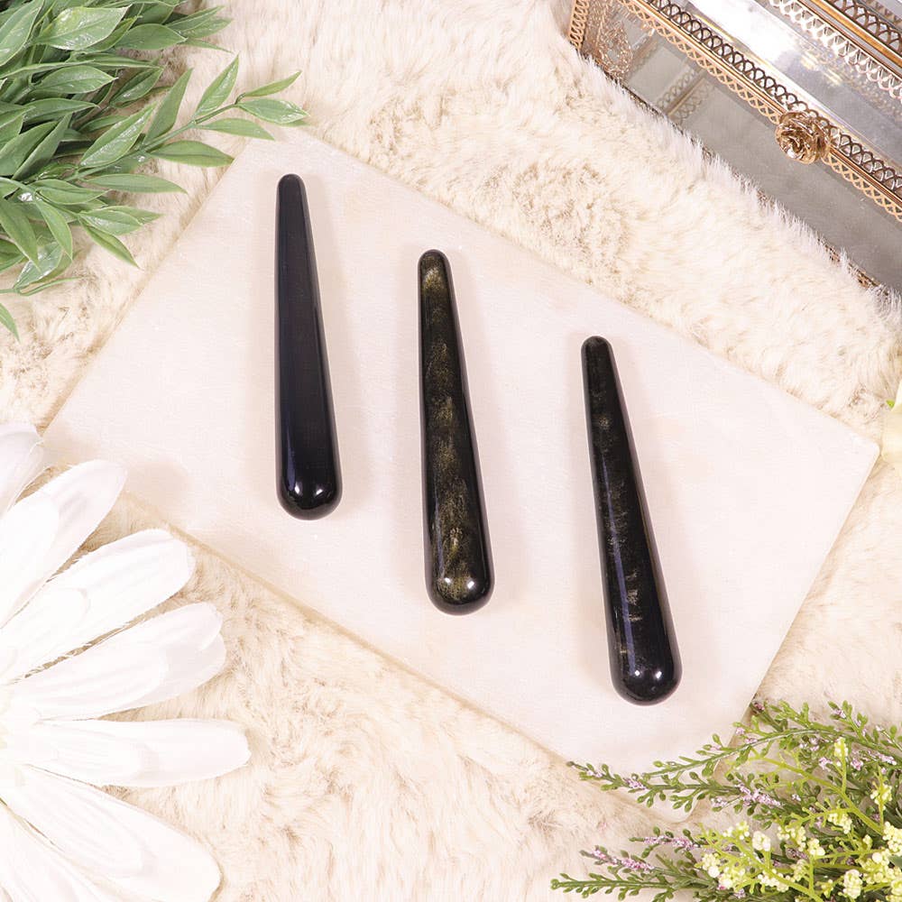 Three golden sheen obsidian wands arranged on a white surface, with a white glove and decorative items in the background.