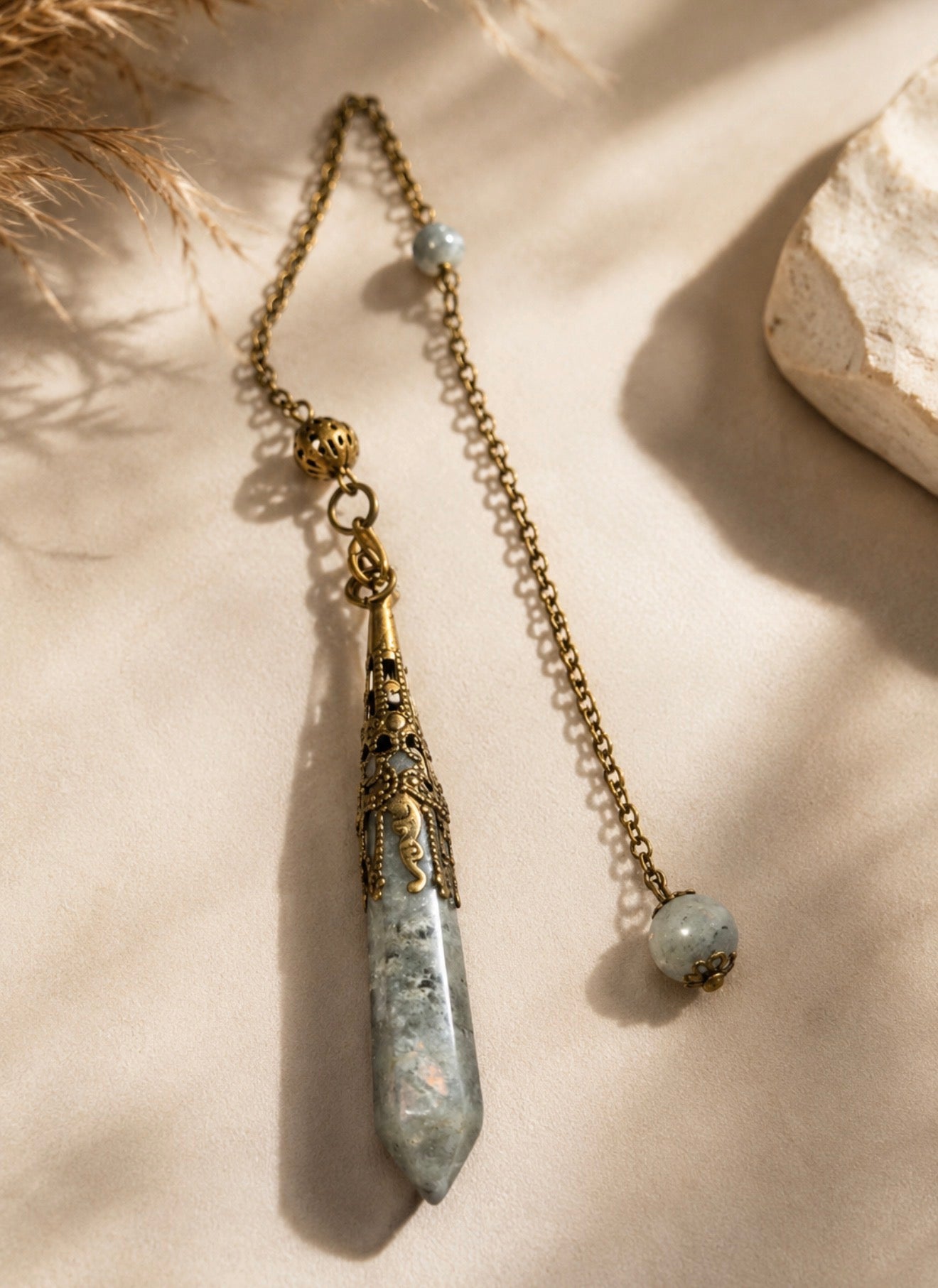 A bohemian-style pendant necklace featuring a large, textured gray and white crystal point pendant with ornate bronze filigree detailing, a matching bronze beaded chain, and dried grass and stone props on a beige background.