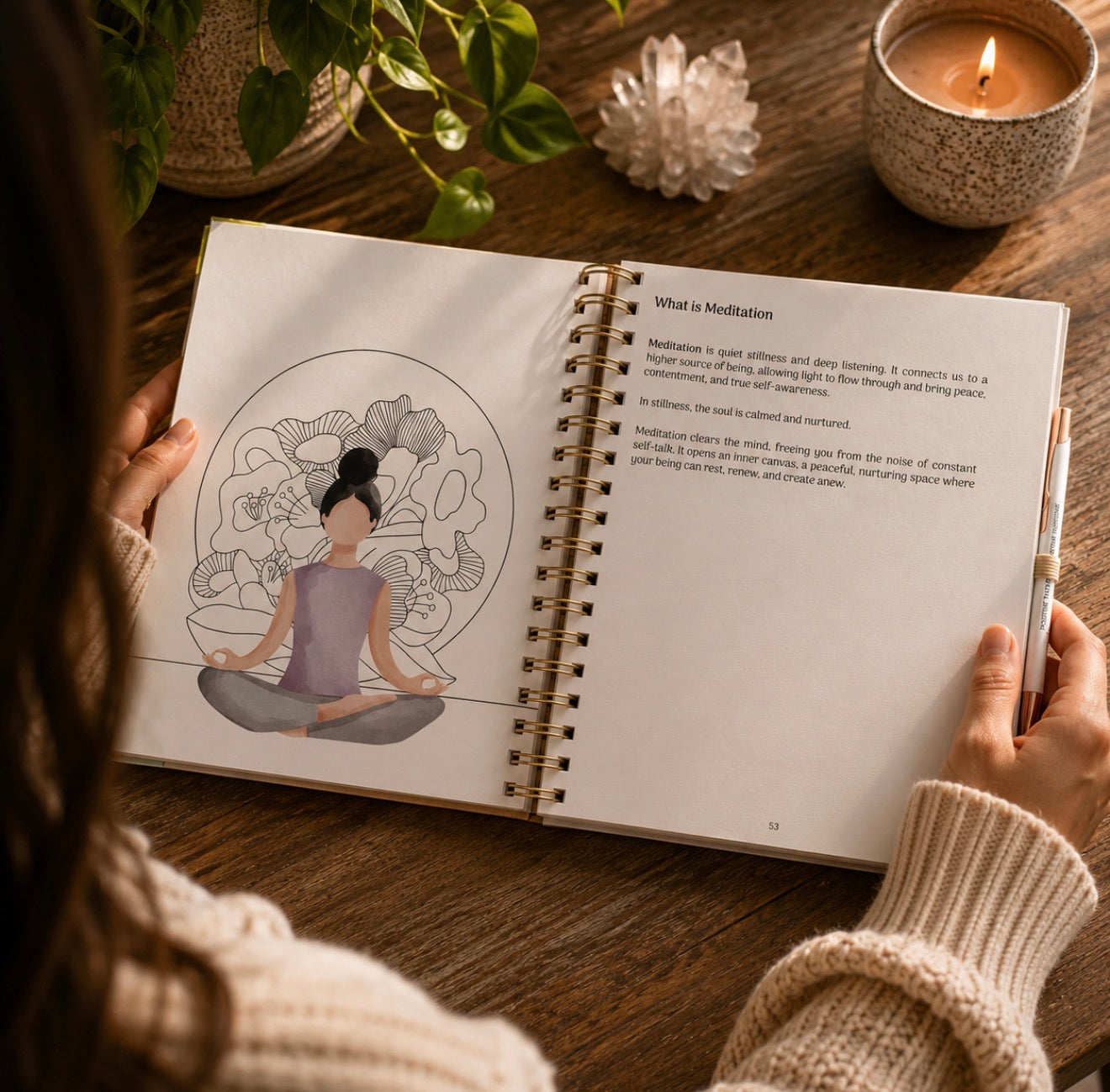 A person holds an open spiral-bound book on a wooden table, showing a page titled "What is Meditation" with text and an illustration of a seated meditating figure. The scene includes a lit candle, a plant, and a crystal in a cozy indoor setting.