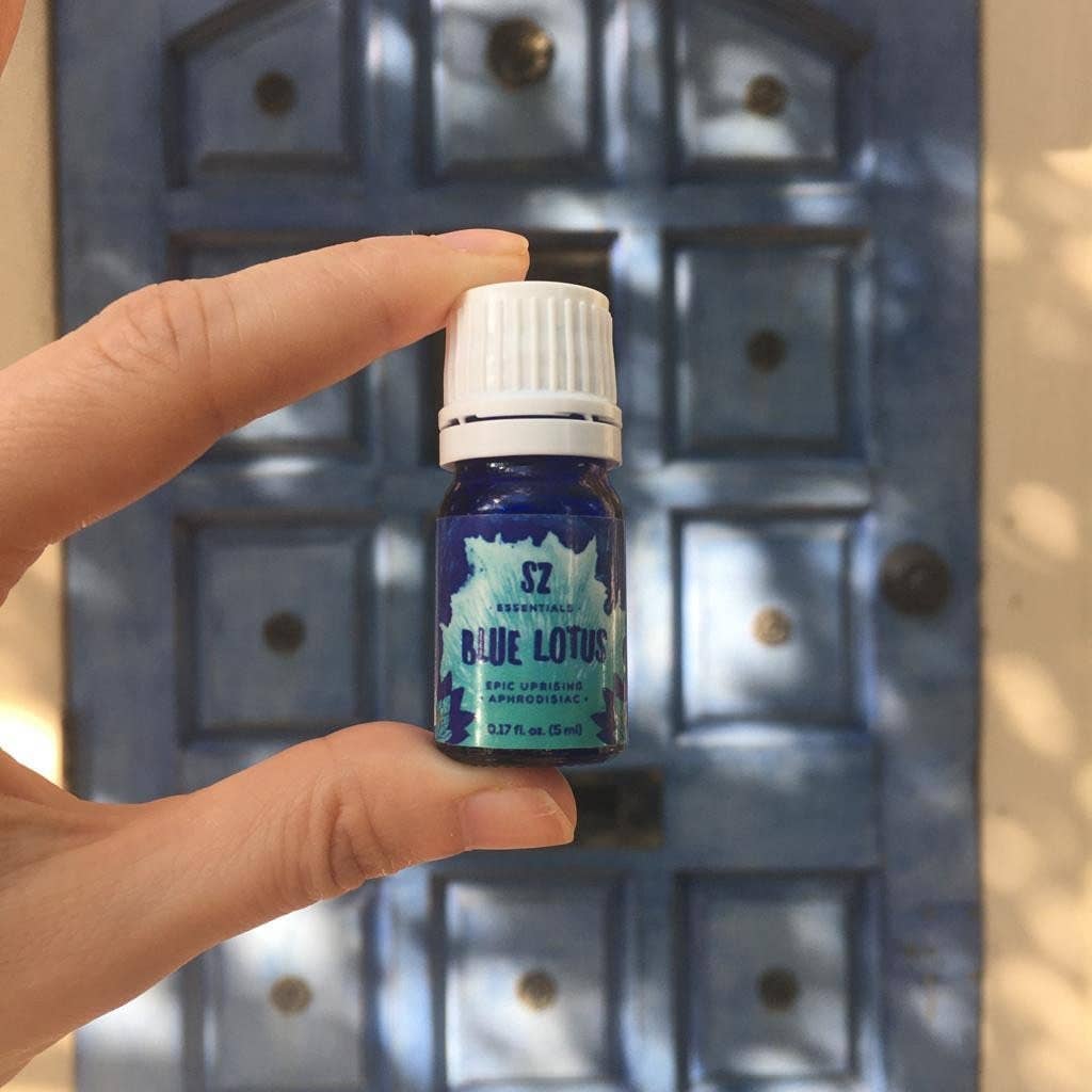 Hand holding a small bottle labeled 'Blue Lotus' against a textured wall background - JOY - Journey Of You - Spiritual, Holistic, Empowering