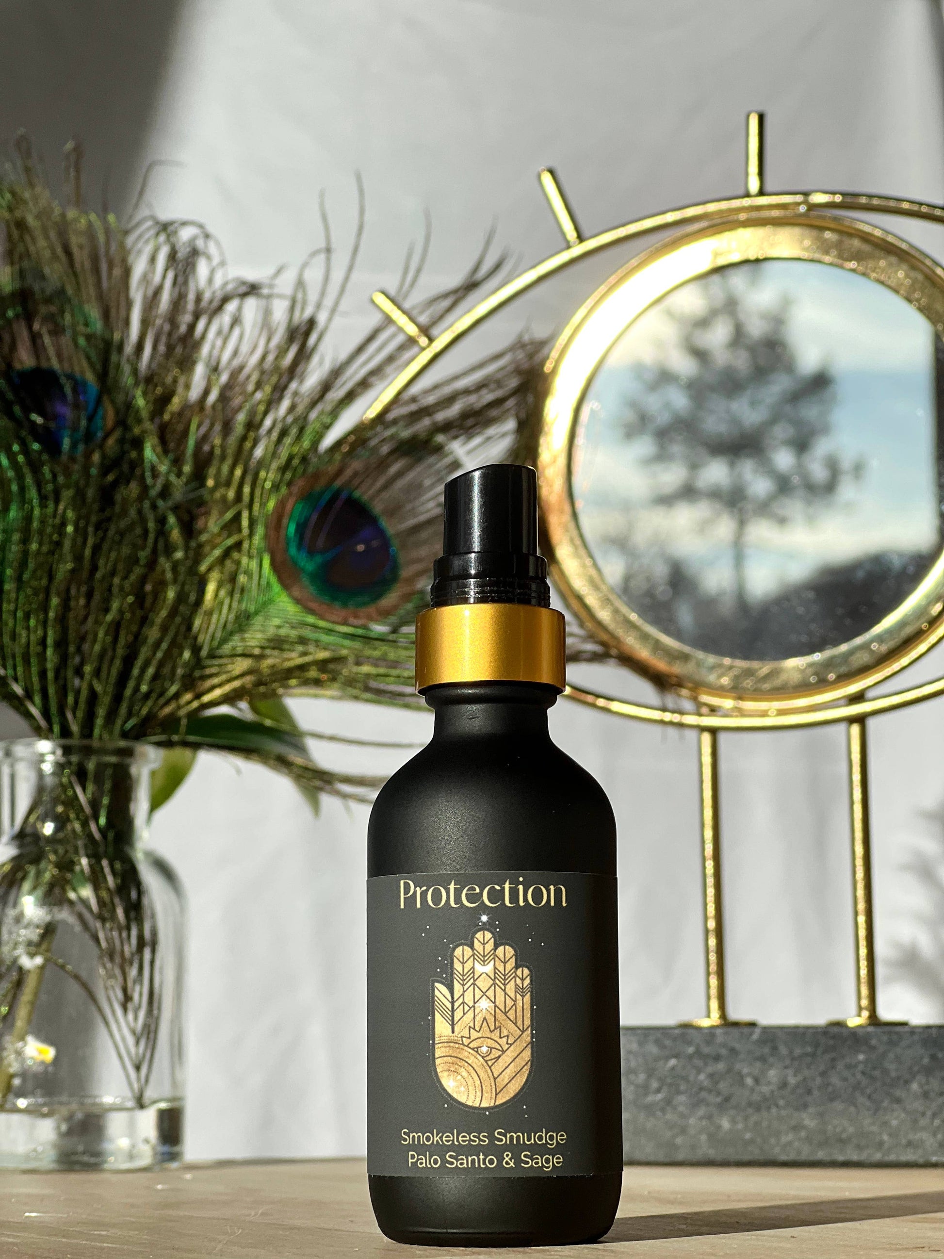 Black bottle labeled 'Protection' with gold accents on a wooden surface, peacock feathers, and decorative mirror in the background.