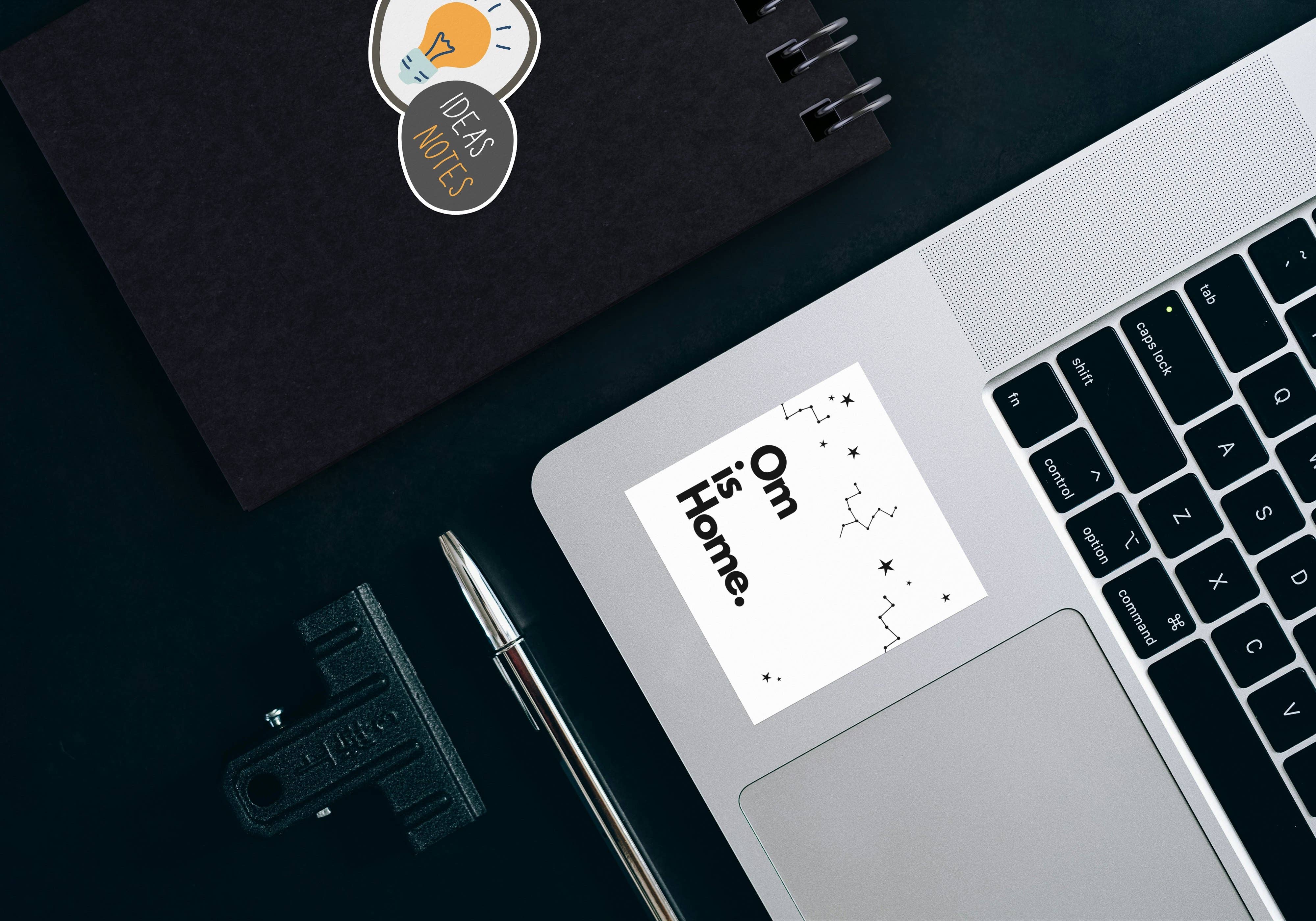 Laptop with a sticker on the touchpad, next to a notebook and pen on a dark surface - JOY - Journey Of You - Spiritual, Holistic, Empowering