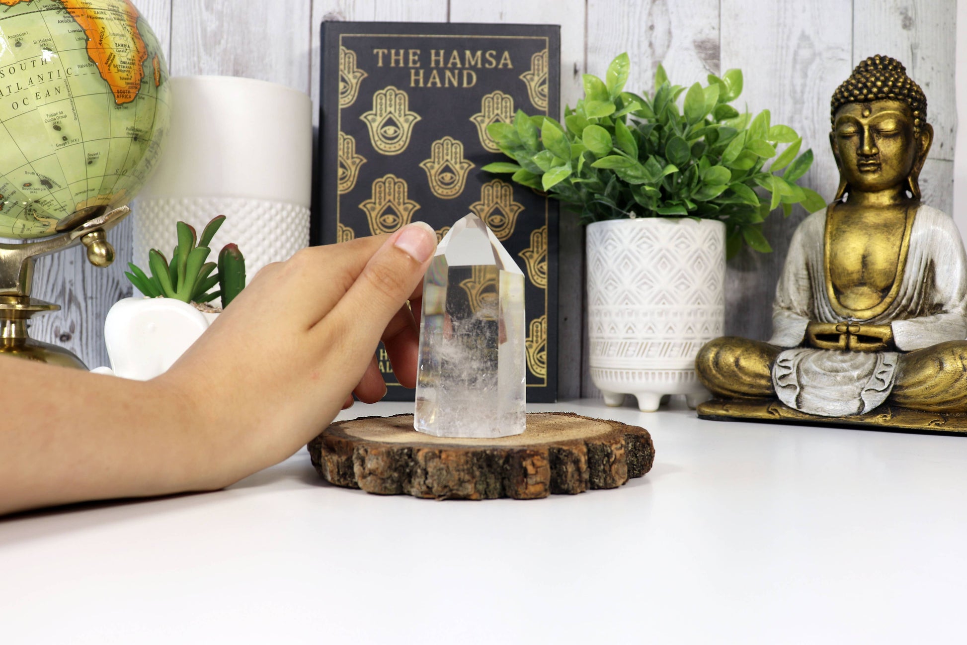 Person holding a crystal tower on a table with decor items including a Buddha statue and a Hamsa hand poster.