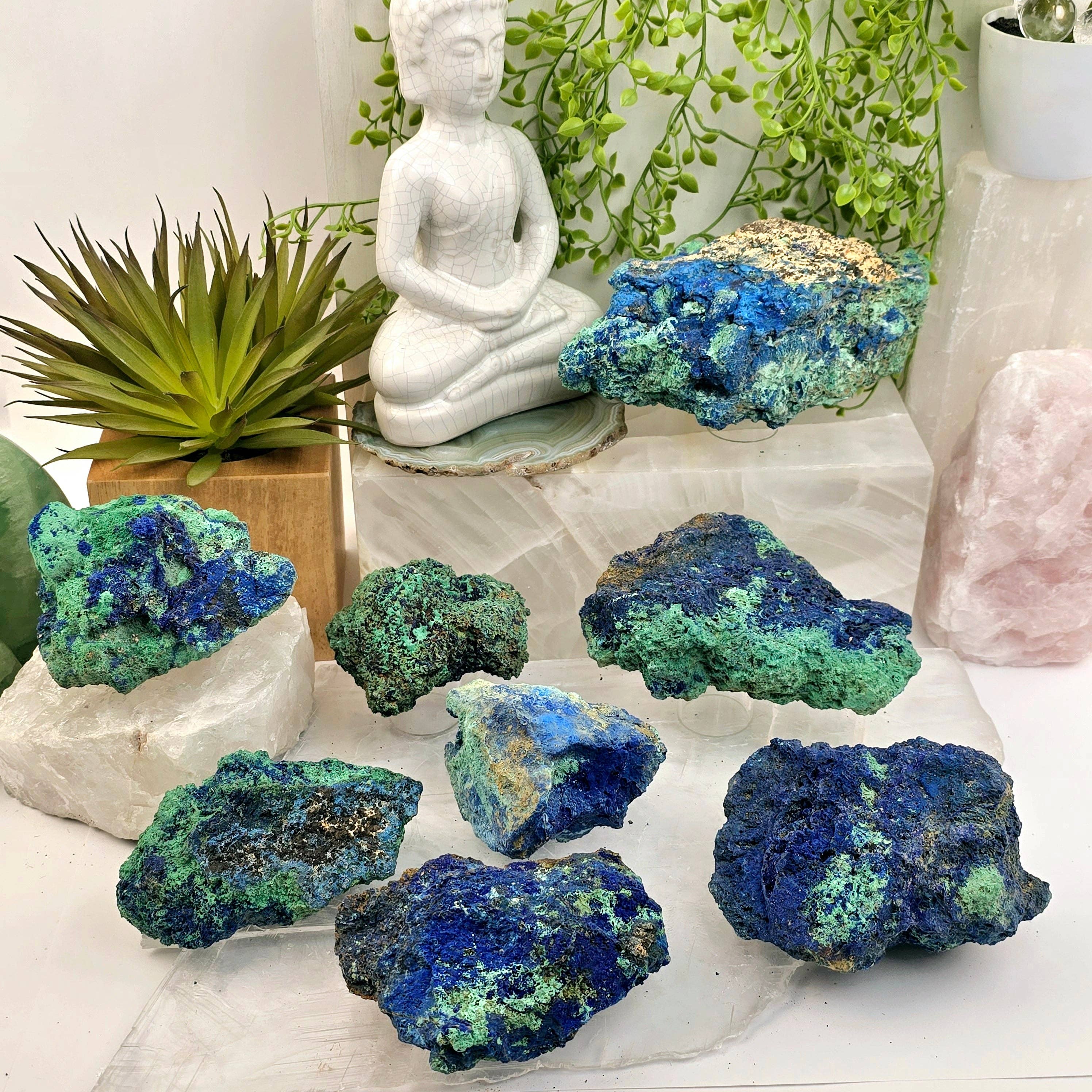 Collection of blue and green crystals on a white surface with a small statue and plants in the background.