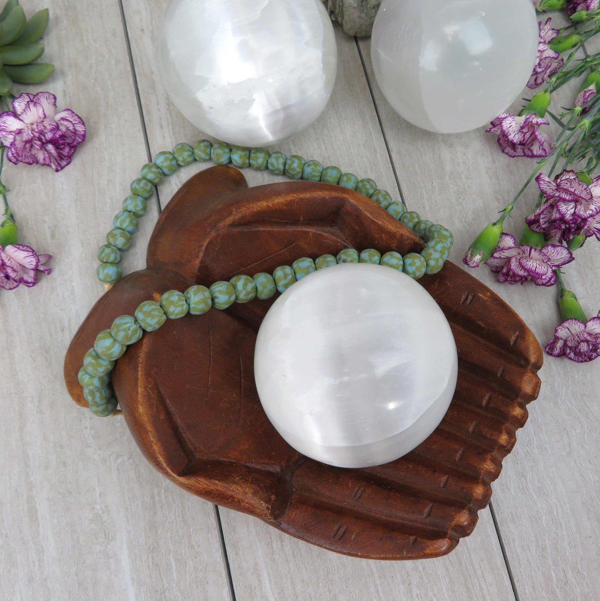 White crystal ball on a wooden hand with green beads, surrounded by flowers on a light wooden surface.