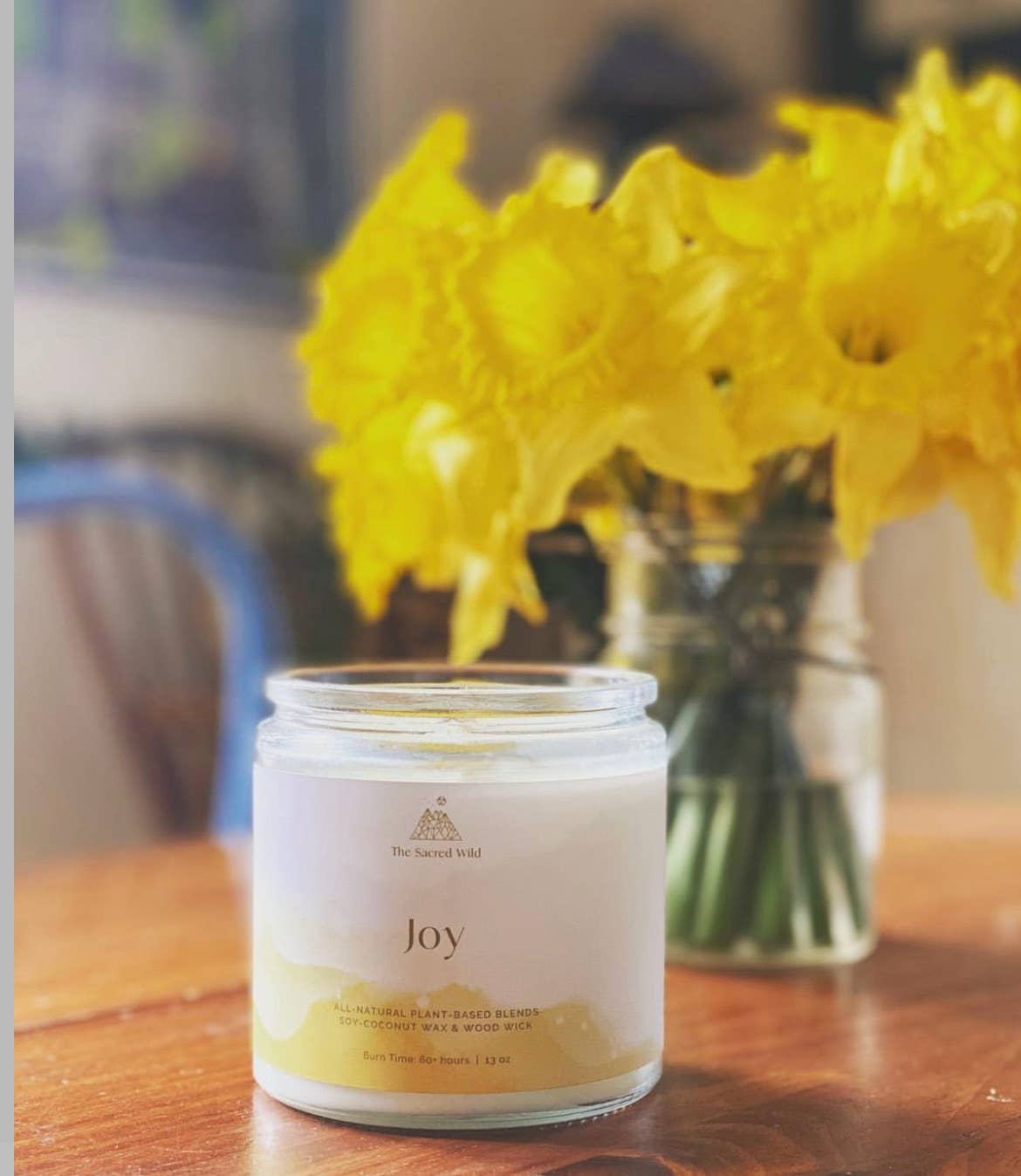 Candle labeled 'Joy' on a table with a vase of yellow flowers in the background
