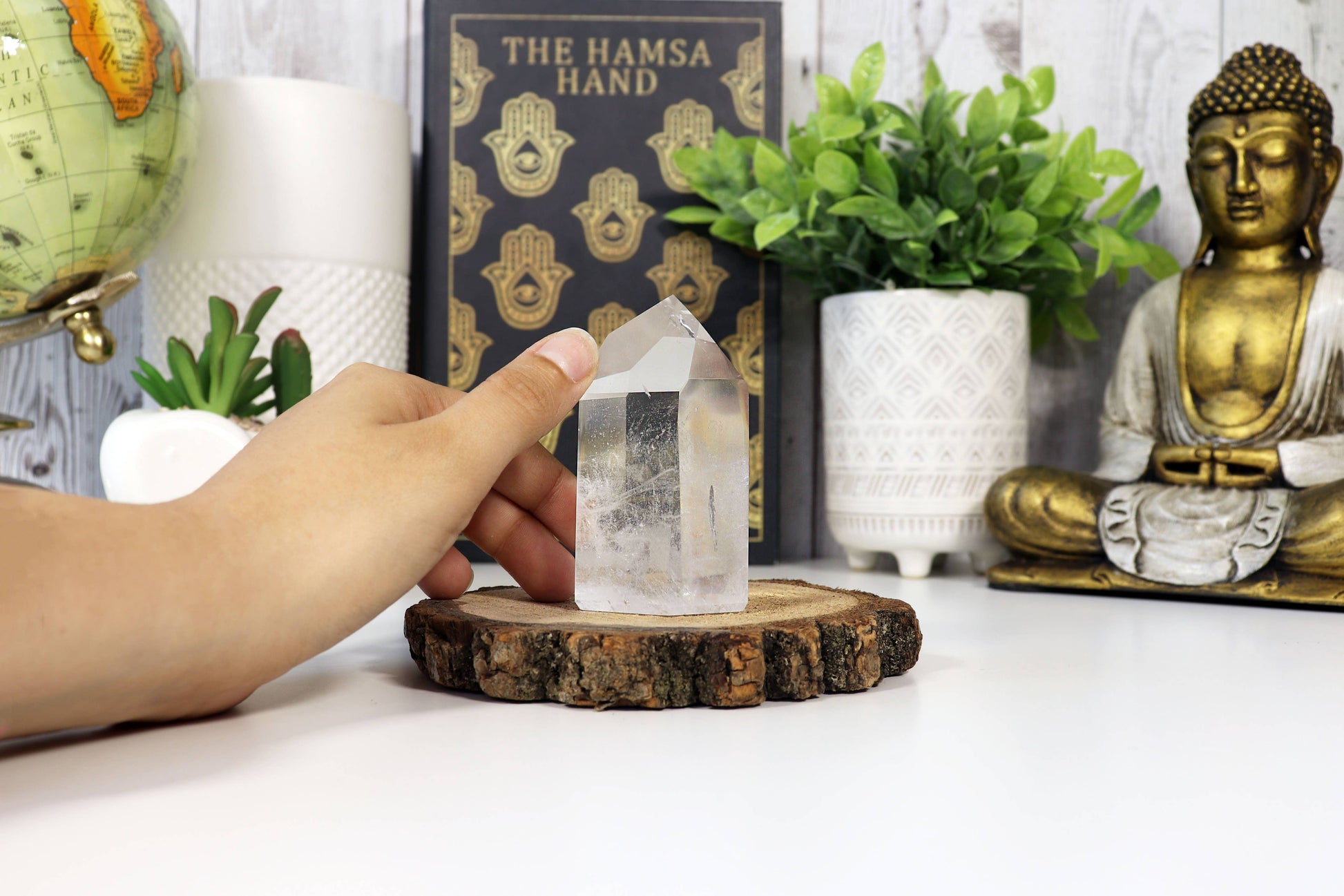 Person holding a crystal on a wooden stand with a Buddha statue, plants, and books in the background.