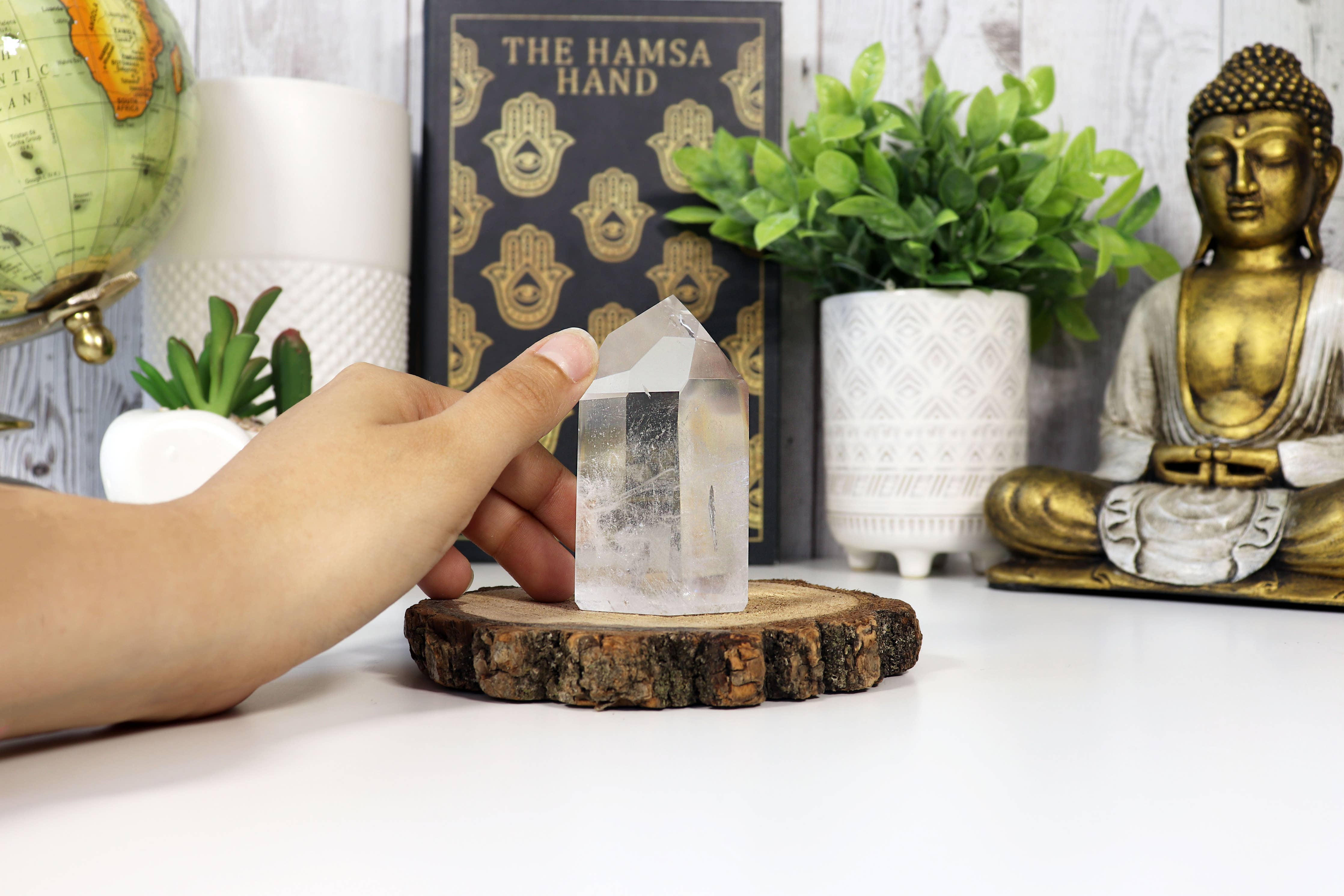 Person holding a crystal on a wooden stand with a Buddha statue, plants, and books in the background.