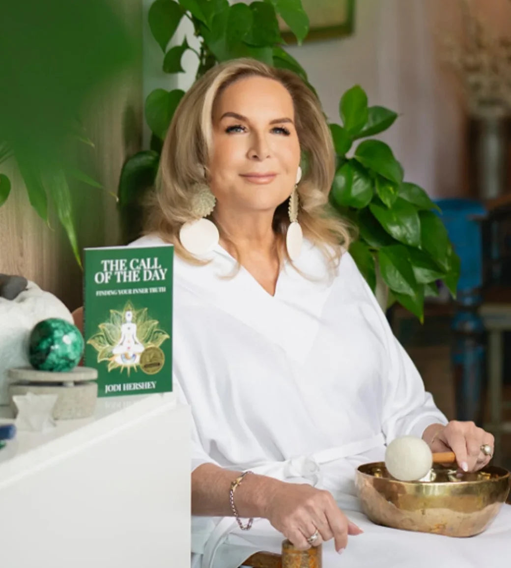 Woman in a white robe holding a brass singing bowl with a book titled 'The Call of the Day' on a table.