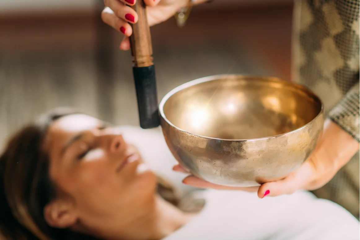 A person holds a polished singing bowl and mallet while another person lies down in the background, suggesting a meditation or therapy session.